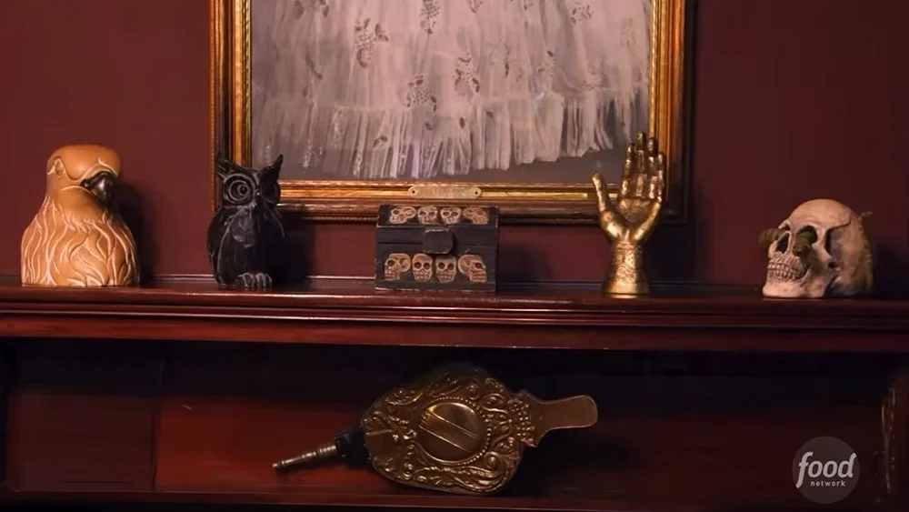 A mantel holding a carved bird, an owl, a skull-covered chest, a golden hand, and a skull, with bellows on the shelf below