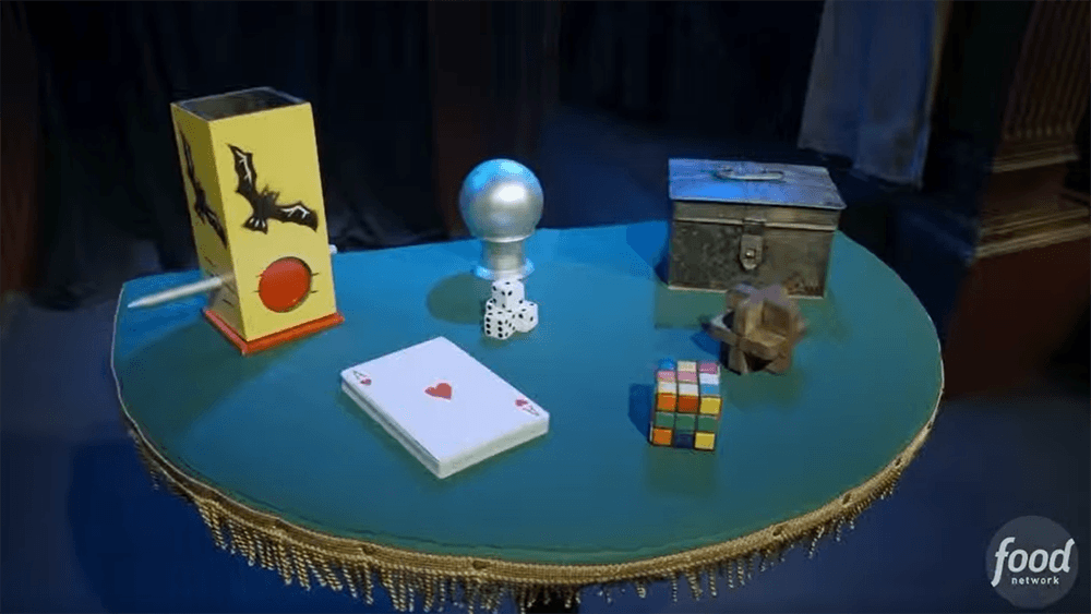 A half-round table with a yellow bat-decorated box, a silver ball, dice, a deck of cards, a metal box, a Rubik’s Cube, and a wooden puzzle
